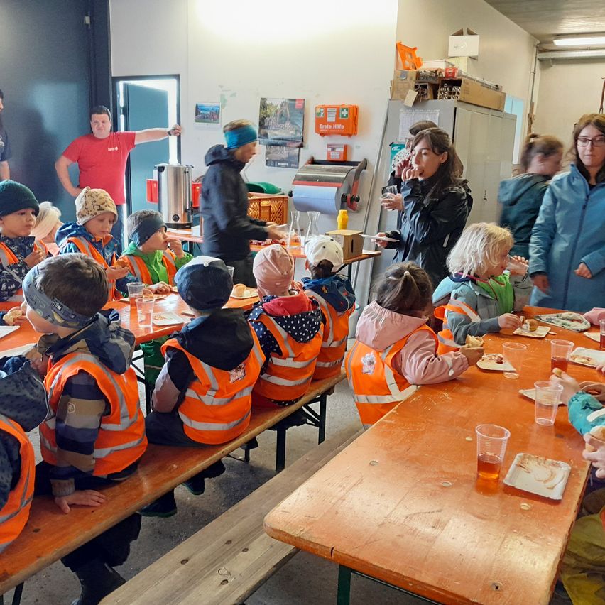 Kinder in orange Westen sitzen um Tische herum und essen. Eine Frau mit Brille steht bei einer Kaffeemaschine, und ein Mann in einem roten Shirt ist im Hintergrund.