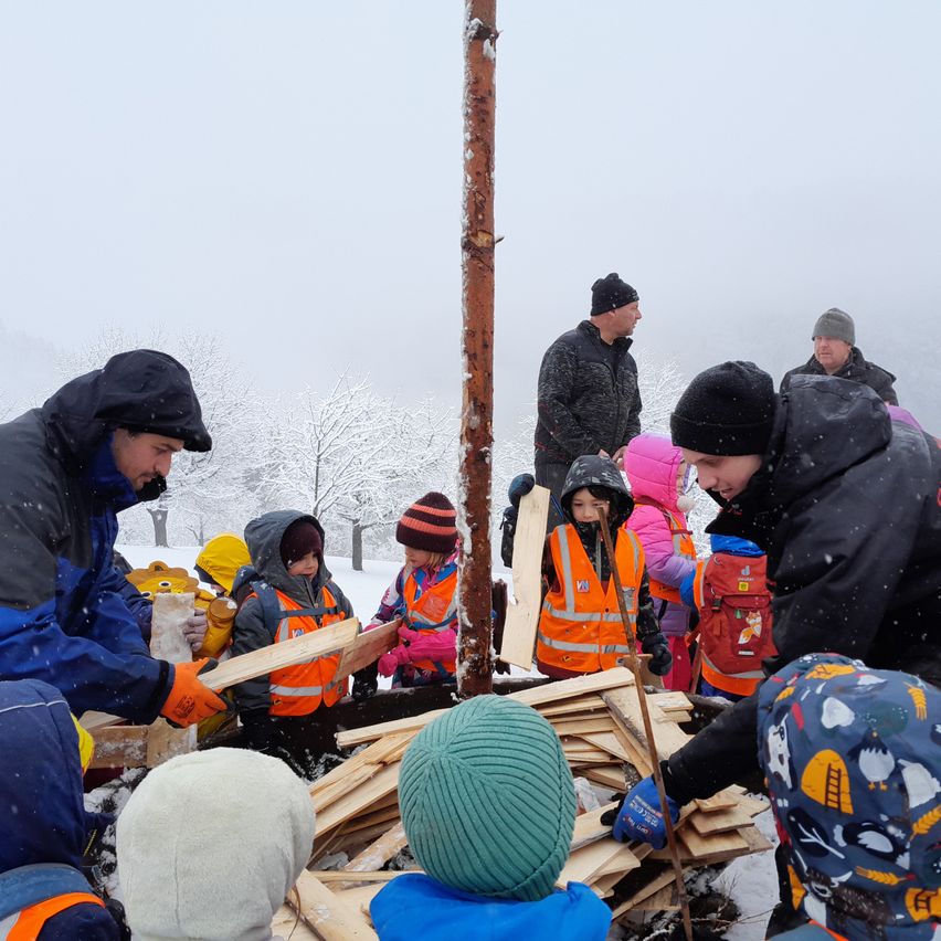 Eine Gruppe von Menschen, darunter Erwachsene und Kinder, ist im Freien im Schnee. Sie tragen Winterkleidung und einige haben Warnwesten an. Sie stehen um einen Holzhaufen herum, wobei zwei Personen Sägen benutzen, um Stücke zu schneiden. Ein Pfosten steht in der Mitte des Haufens. Im Hintergrund sind Bäume zu sehen.