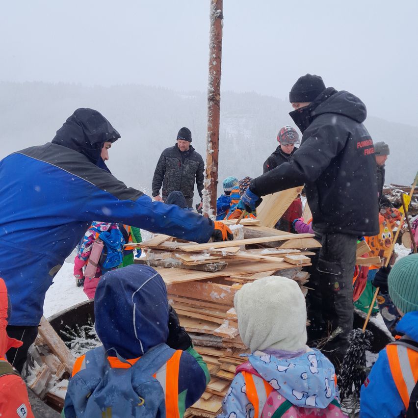 Erwachsene und Kinder ziehen Winterjacken und Mützen an, während sie Holz in einem verschneiten Außenbereich stapeln. Einige tragen Rucksäcke, und ein Kind hält eine Holzplanke. Im Hintergrund steht ein Mann in der Nähe eines Pfostens, und Berge sind sichtbar.