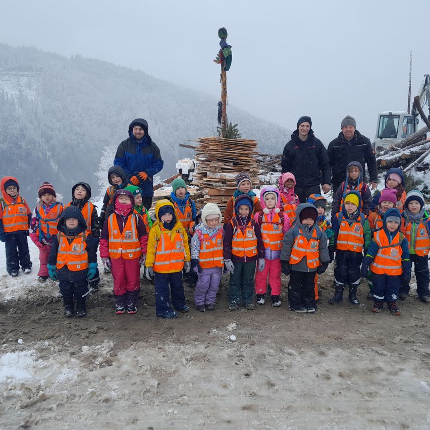 Eine Gruppe von Kindern in orangefarbenen Westen posiert für ein Foto bei verschneiten Bedingungen mit Erwachsenen, Bergen und Maschinen im Hintergrund.