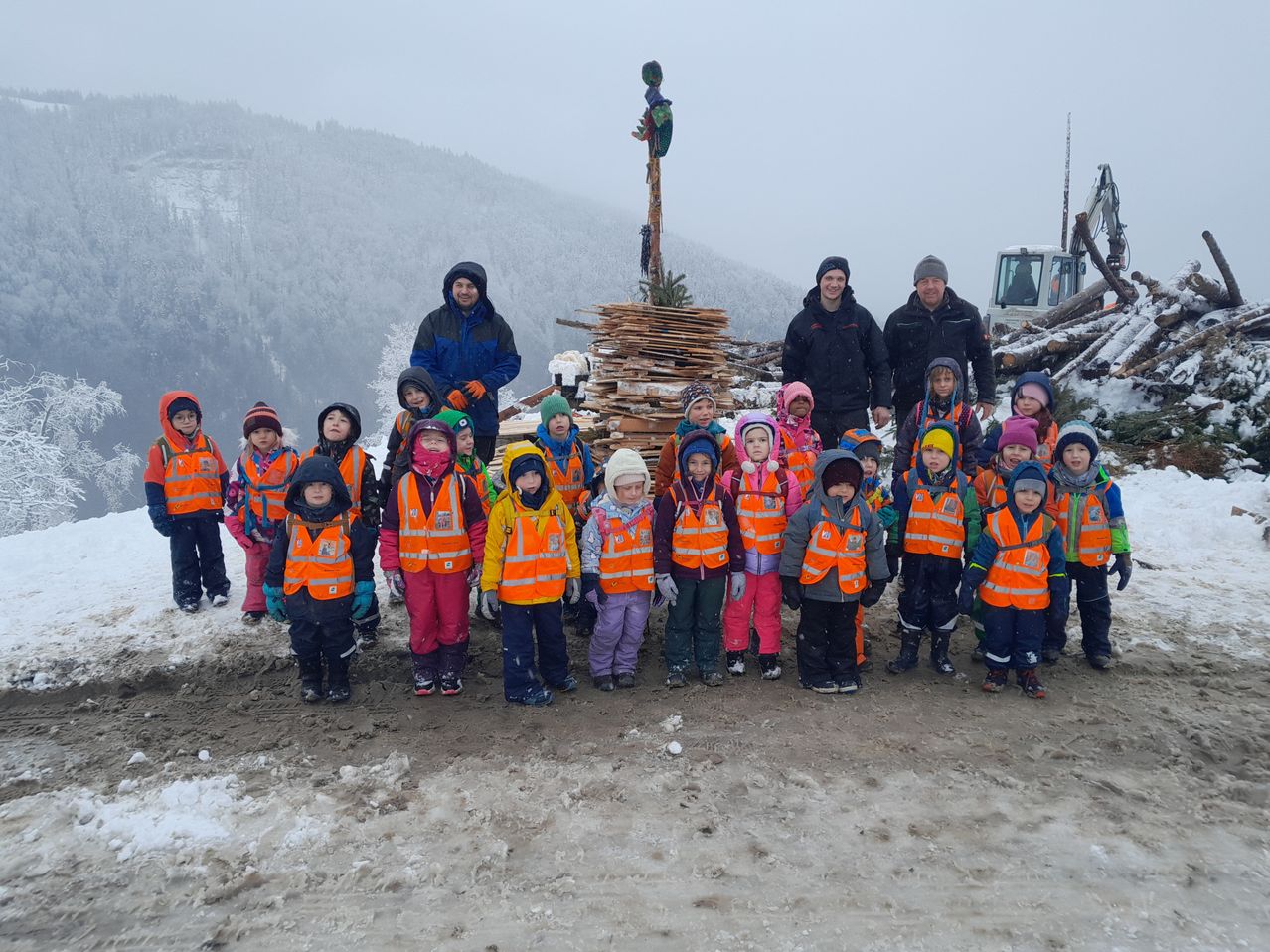 Eine Gruppe von Kindern in orangefarbenen Westen posiert für ein Foto bei verschneiten Bedingungen mit Erwachsenen, Bergen und Maschinen im Hintergrund.