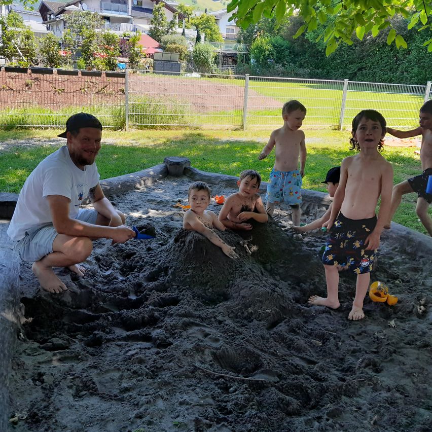 Ein Mann und mehrere Kinder spielen in einem Sandkasten. Sie sind von einem Zaun umgeben, mit Häusern und Bäumen im Hintergrund.