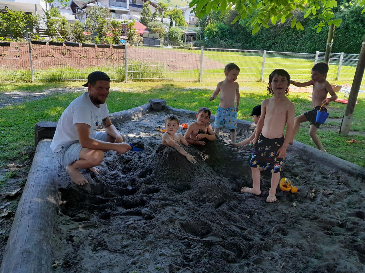 Ein Mann und mehrere Kinder spielen in einem Sandkasten. Sie sind von einem Zaun umgeben, mit Häusern und Bäumen im Hintergrund.