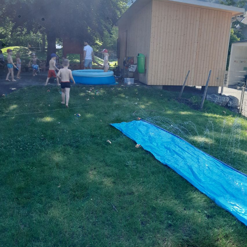 Kinder spielen im Garten mit einem Wasserrutsche, einem aufblasbaren Pool und einem Holzhaus. Im Hintergrund befinden sich Bäume und ein Zaun.