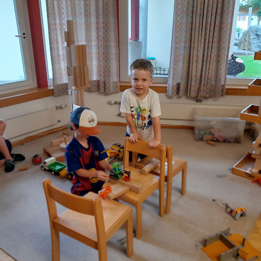 Zwei Jungen spielen mit Holzspielzeug in einem Raum. Ein Junge steht auf einem Stuhl, während der andere sitzt. Verschiedene Spielzeuge sind herum verstreut.