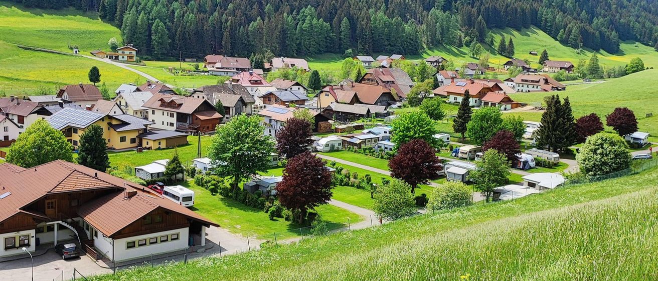 Luftaufnahme eines ländlichen Dorfes mit Häusern, Bäumen und einer Straße, umgeben von grünen Feldern und Bergen im Hintergrund.