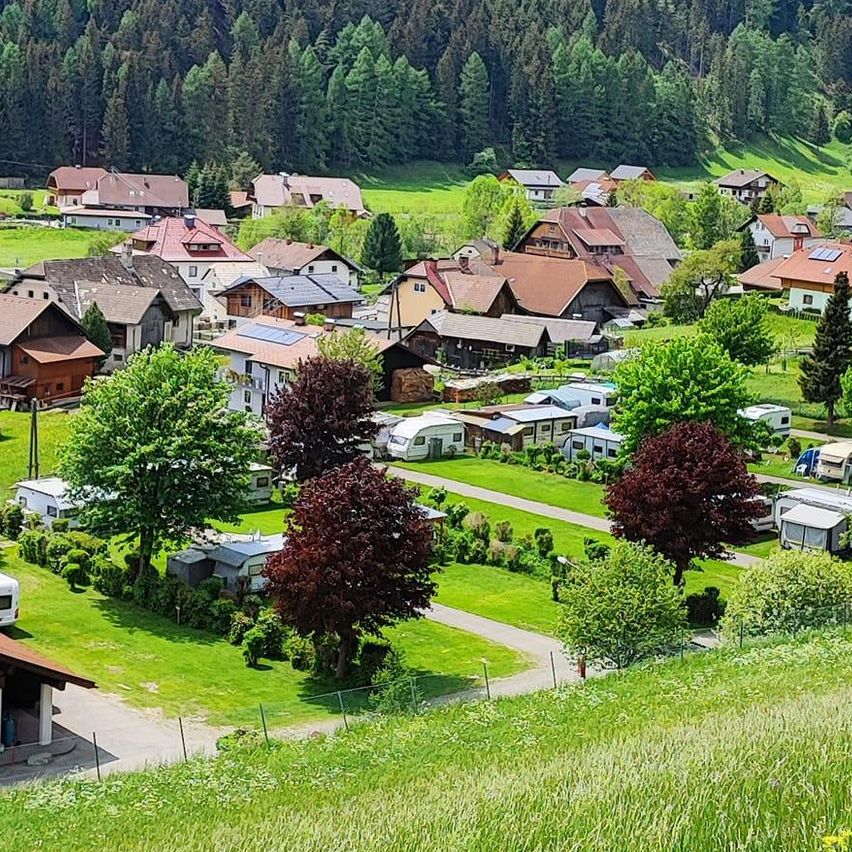 Luftaufnahme eines ländlichen Dorfes mit Häusern, Bäumen und einer Straße, umgeben von grünen Feldern und Bergen im Hintergrund.