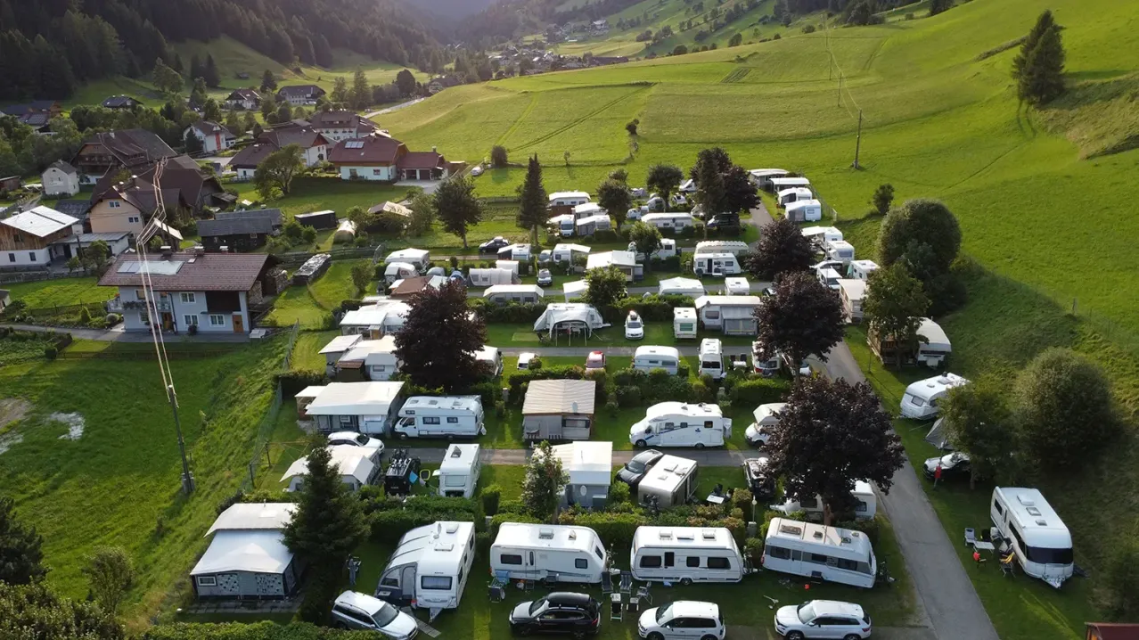 Eine Luftaufnahme eines ländlichen Campingplatzes mit zahlreichen weißen Wohnmobilen, einige mit Markisen, auf einem Rasenfeld geparkt. Bäume und Büsche sind verstreut. Gebäude sind im Hintergrund sichtbar.