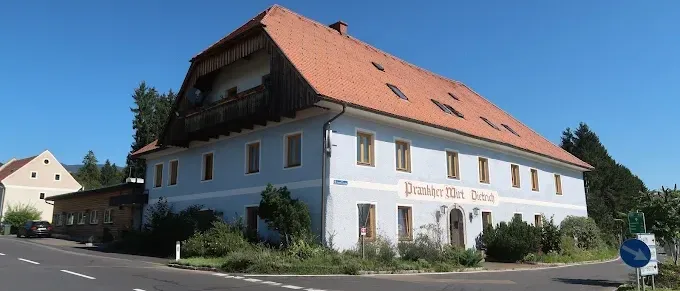 Ein weißes Gebäude mit rotem Dach und mehreren Fenstern befindet sich an einer Straßenecke mit viel Grün und einem klaren blauen Himmel.