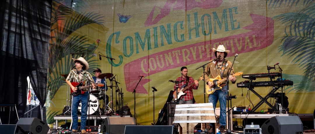 Eine Country-Band spielt auf der Bühne mit einem Hintergrund, der 'Coming Home Country Festival' besagt. Der Leadsänger hält ein Mikrofon und eine Gitarre, während andere Gitarre, Schlagzeug und Keyboard spielen.