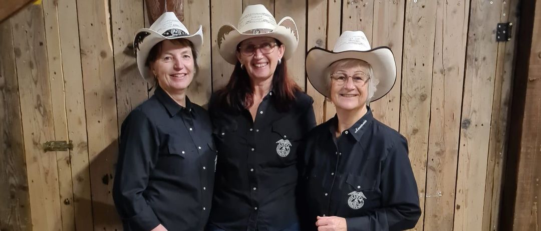 Drei Frauen in Cowboyhüten und schwarzen Hemden stehen nebeneinander vor einer Holzwand. Sie alle lächeln und scheinen für ein Foto zu posieren.