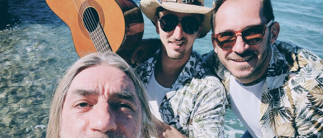 Drei Männer stehen im flachen Wasser am Strand und lächeln für ein Selfie. Der Mann in der Mitte hält eine Gitarre, während die anderen Sonnenbrillen und Hüte tragen.