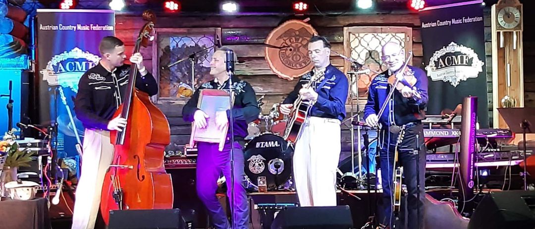 Vier Musiker spielen auf einer Bühne Cello, Gitarre, Geige und Keyboard. Im Hintergrund steht eine Blumendekoration und Scheinwerfer. Ein Banner der Austrian Country Music Federation ist sichtbar.