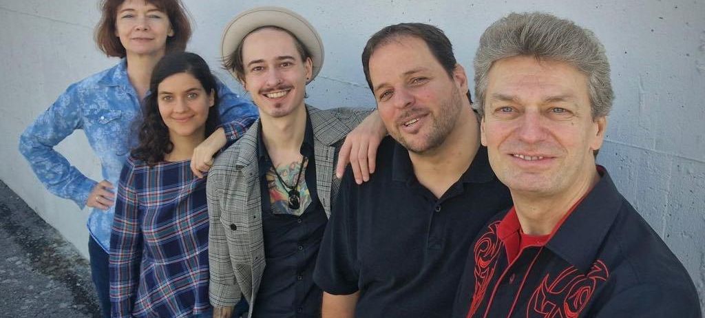 Fünf Personen posieren für ein Foto vor einer weißen Wand. Zwei Frauen stehen links, eine mit einem karierten Shirt und die andere mit einer Jeansjacke. In der Mitte lächelt ein Mann mit einem Hut. Die beiden anderen Männer tragen schwarze Hemden, einer mit einem roten Kragen.