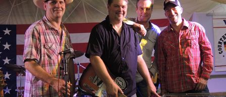 Vier Männer stehen lächelnd auf einer Bühne. Einer hält eine Gitarre, ein anderer spielt Keyboard. Zwei weitere stehen dahinter, einer mit einem Mikrofon. Dahinter ein rot-weiß gestreifter Hintergrund.