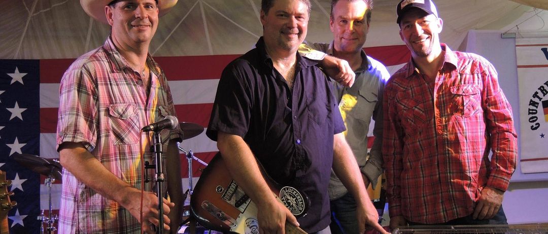 Vier Männer stehen lächelnd auf einer Bühne. Einer hält eine Gitarre, ein anderer spielt Keyboard. Zwei weitere stehen dahinter, einer mit einem Mikrofon. Dahinter ein rot-weiß gestreifter Hintergrund.