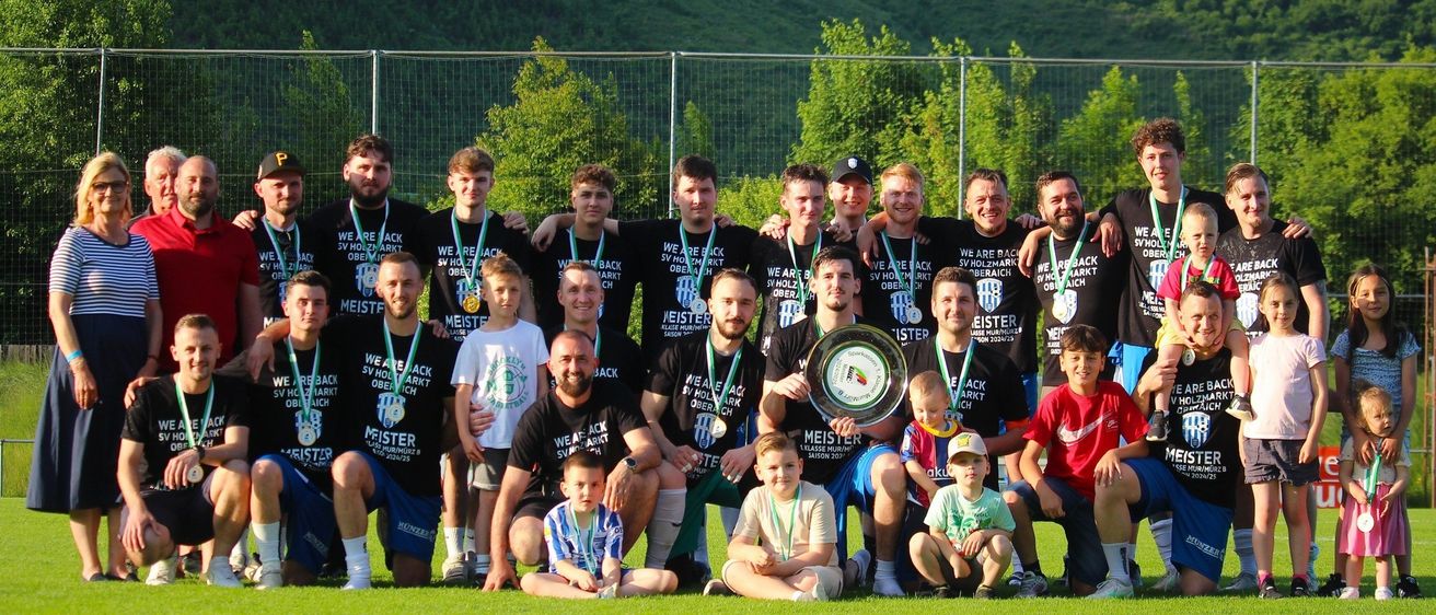 Eine Fußballmannschaft posiert für ein Gruppenfoto mit einem Pokal auf einem Feld, umgeben von einem Metallzaun und Grünflächen im Hintergrund.