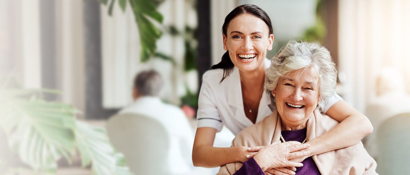 Eine lächelnde ältere Frau in einem lila Pullover wird von einer Krankenschwester umarmt, beide stehen eng beieinander. Ein Mann sitzt auf einem Stuhl im Hintergrund.