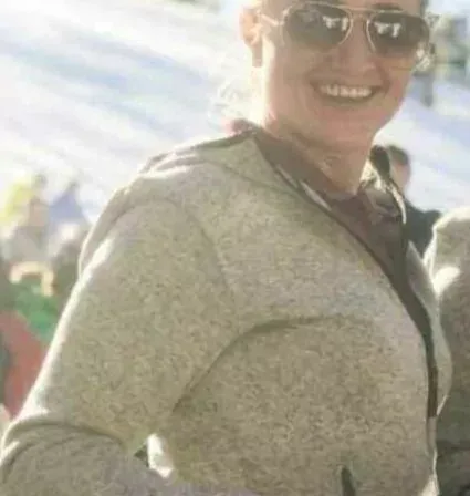 Eine Frau mit Sonnenbrille hält eine Schüssel und lächelt in einer verschneiten Umgebung mit Leuten im Hintergrund.