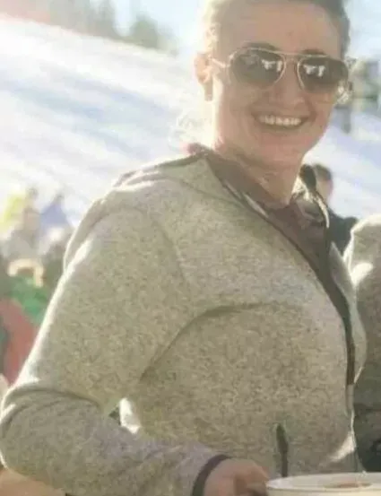 Eine Frau mit Sonnenbrille hält eine Schüssel und lächelt in einer verschneiten Umgebung mit Leuten im Hintergrund.