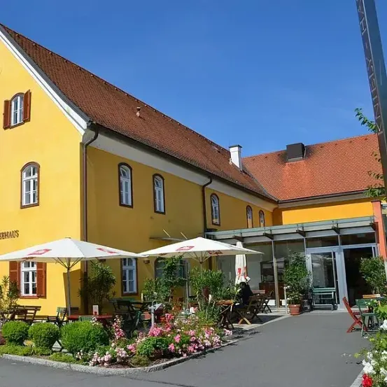 Ein gelbes Gebäude mit mehreren Fenstern und einem roten Dach. Eine Person sitzt unter einem Regenschirm an einem Tisch draußen. Der vordere Bereich hat einen Garten mit rosa und weißen Blumen.