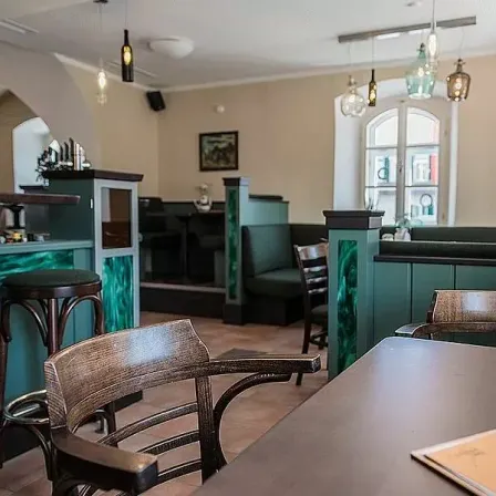 Ein Restaurant mit Holzstühlen und Tischen, einer grünen Bartheke und Pendelleuchten. Die Wände sind beige, und ein Bilderrahmen ist an der Wand montiert.