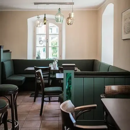 Das Innere eines Restaurants mit einer Bank, Tischen und Stühlen. Die Decke hat hängende Lampen und einen Bilderrahmen an der Wand. Es gibt ein Fenster mit Blick nach draußen.
