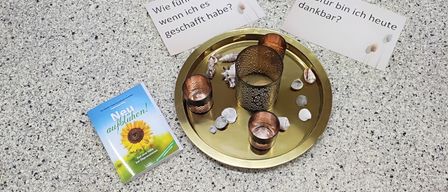 A gold tray with three candles and some stones sits on a table. A small book titled 'New Beginnings' is nearby. Two cards with German text are also present.