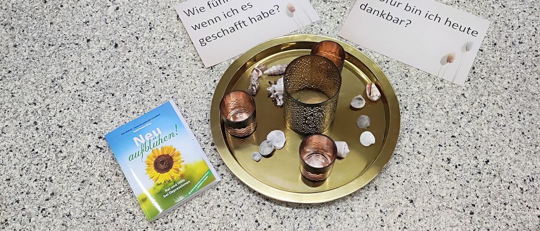 A gold tray with three candles and some stones sits on a table. A small book titled 'New Beginnings' is nearby. Two cards with German text are also present.