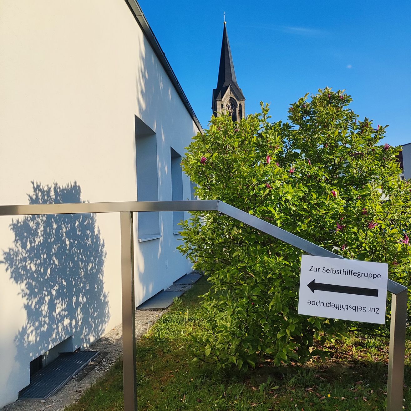 Ein weisses Gebäude mit einem Schatten an der Wand, einem Metallgeländer und einem Busch davor. Ein Schild weist auf eine Selbsthilfegruppe hin. Dahinter steht eine Kirche mit einem Turm.