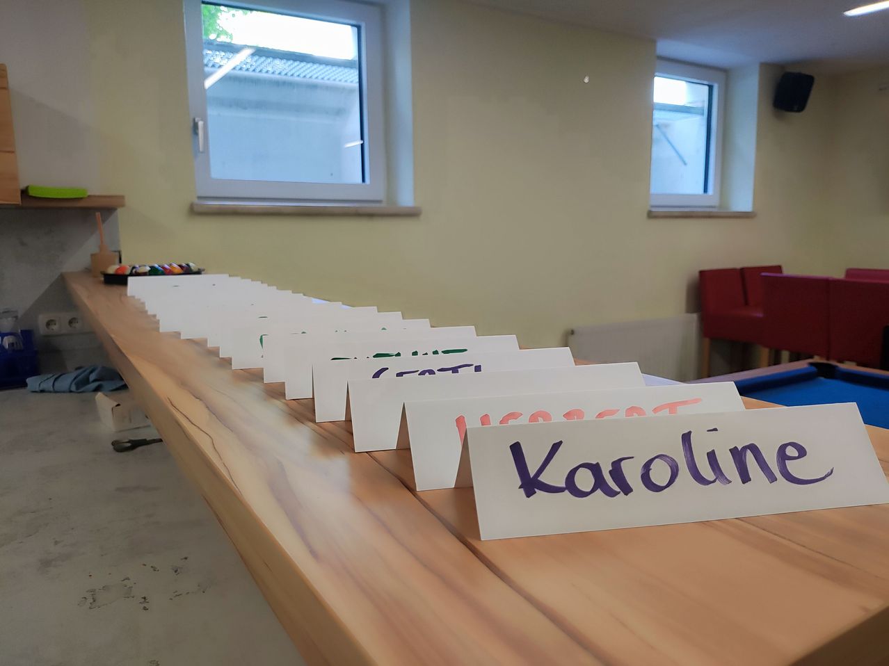 A long wooden table with multiple name tags on it, one of which reads 'Karoline'. The room has two windows and a red chair.