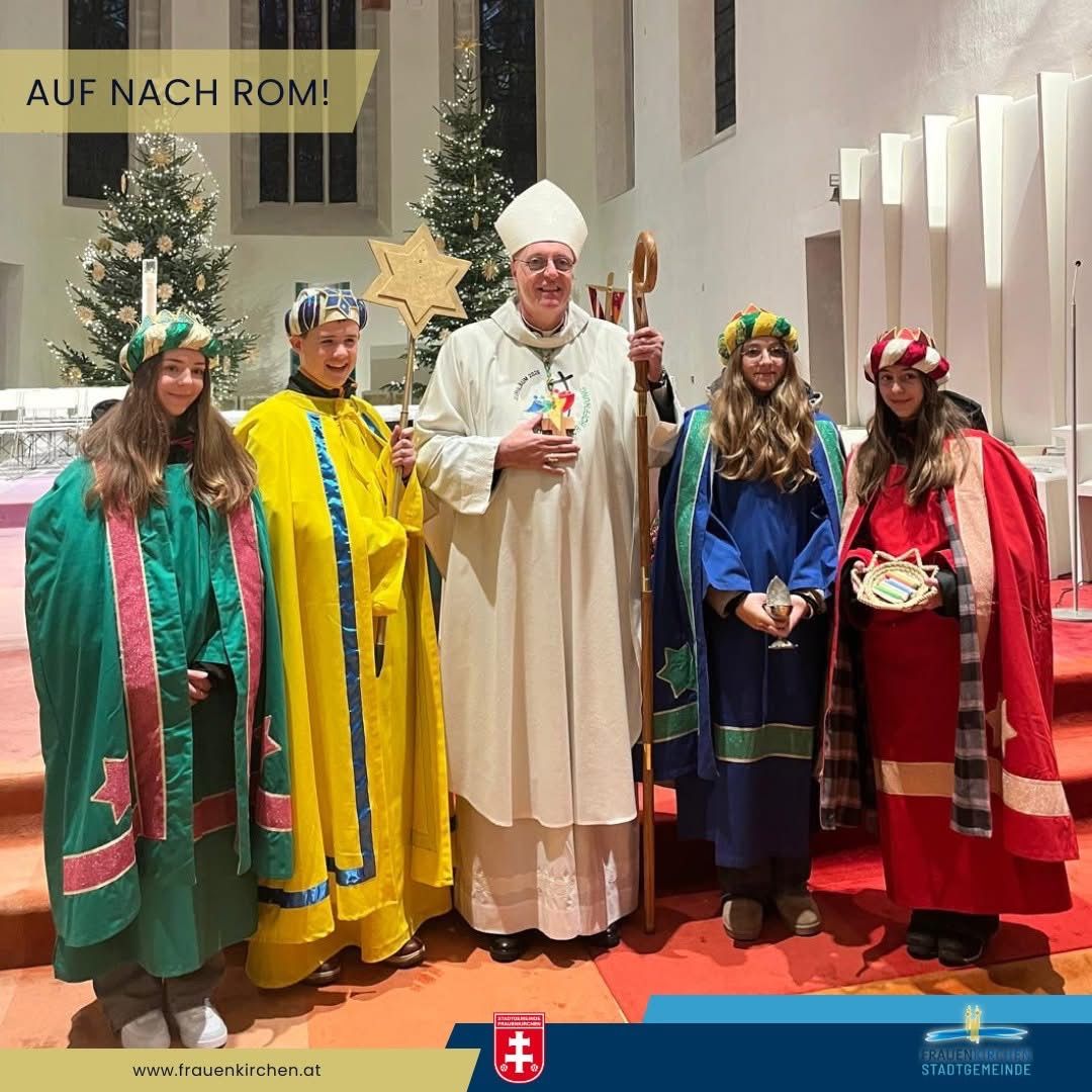 Ein Priester in Weiß steht mit vier jungen Leuten in farbenfrohen Kostümen, die einen Stern und einen Stab halten, vor einem Weihnachtsbaum und einer Kircheninneneinrichtung.