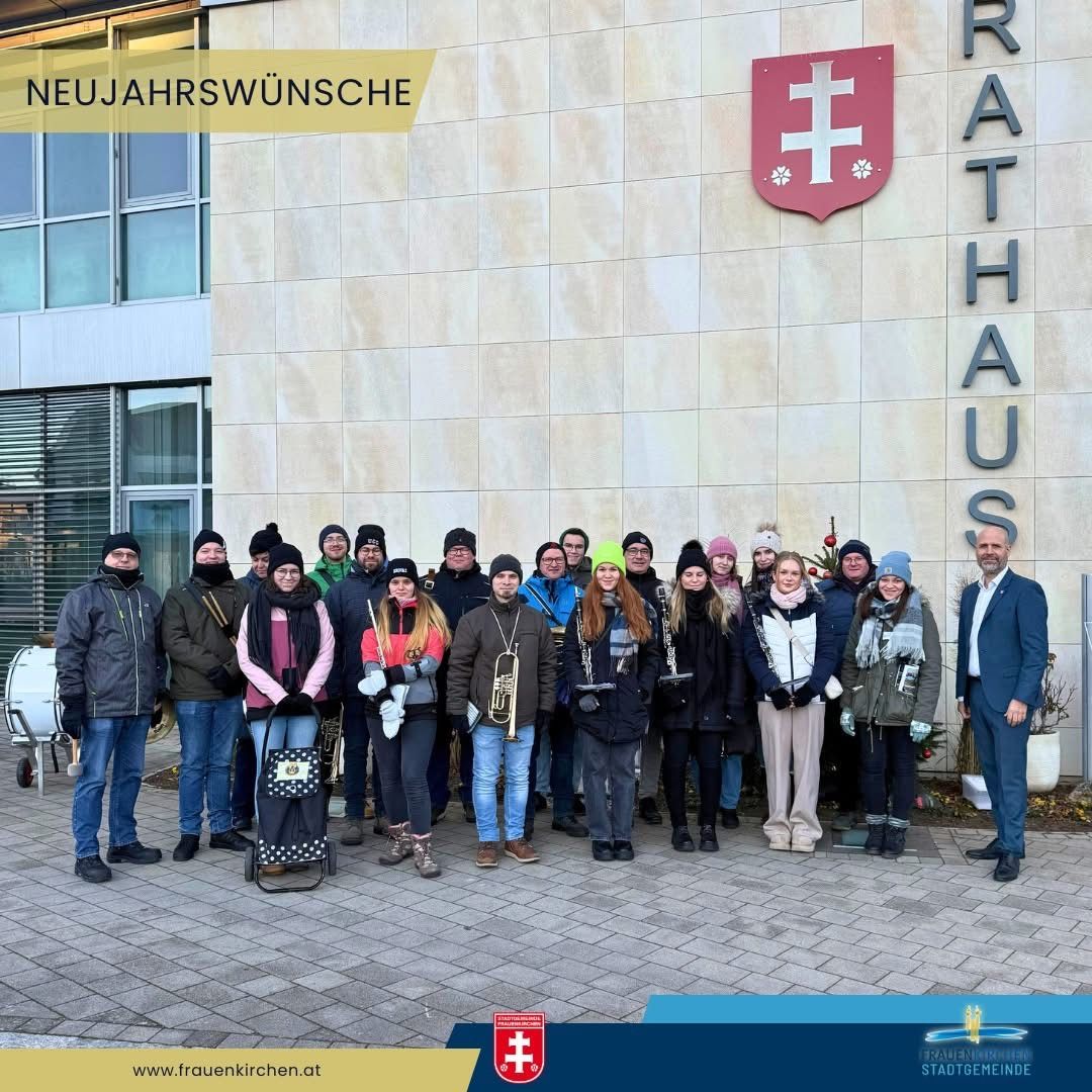 Eine Gruppe von Menschen posiert für ein Foto vor einem Gebäude mit einem Schild, das 'Rathaus' steht. Sie sind alle für kaltes Wetter gekleidet. Einige halten Musikinstrumente. Ein Trommel ist auf der linken Seite platziert. Es gibt einen Wasserzeichen in der unteren linken Ecke.