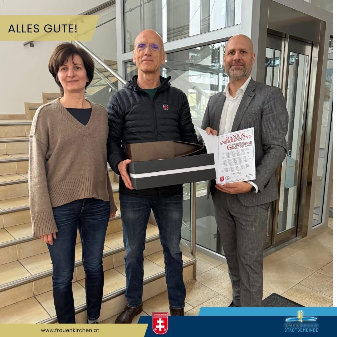 Drei Personen stehen in einem modernen Gebäude. Ein Mann hält ein Zertifikat und eine Box und lächelt. Eine Frau in einem Pullover und Jeans lächelt neben ihm. Ein Mann in einem Anzug schaut auf das Zertifikat. Hinter ihnen befindet sich eine Treppe und eine Glastür. Der Text 'ALLES GUTE!' ist oben.