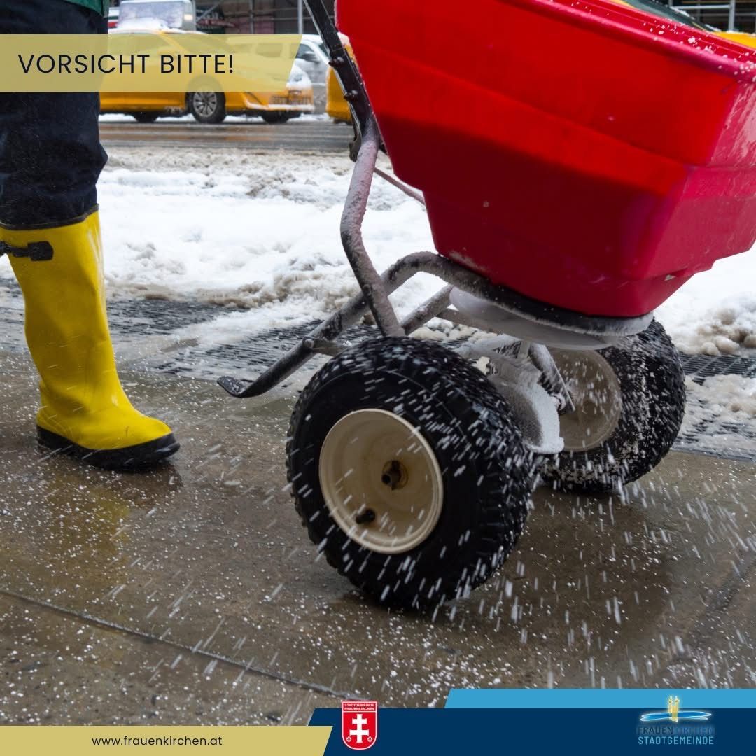 Eine Person in gelben Gummistiefeln steht auf einem nassen Bürgersteig und schiebt einen roten Streuwagen, der Schnee und Wasser verstreut. Im Hintergrund fährt ein Auto vorbei. Oben sind die Worte 'VORSICHT BITTE!' zu sehen.