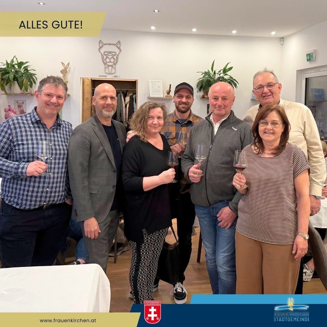 A group of people stand together in a room holding wine glasses. The man on the left wears a plaid shirt, and the woman in the center wears a black shirt. Above them is a yellow banner that reads 'ALLES GUSTE!'.