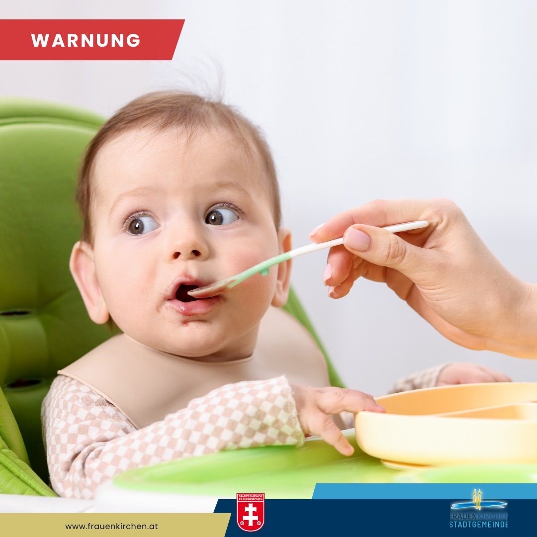 Ein Baby sitzt in einem Hochstuhl und wird von einer Person mit Löffel gefüttert. Das Baby trägt ein Lätzchen und der Löffel ist grün. Das Bild hat einen Wasserzeichen von 'www.frauenkirchen.at'.