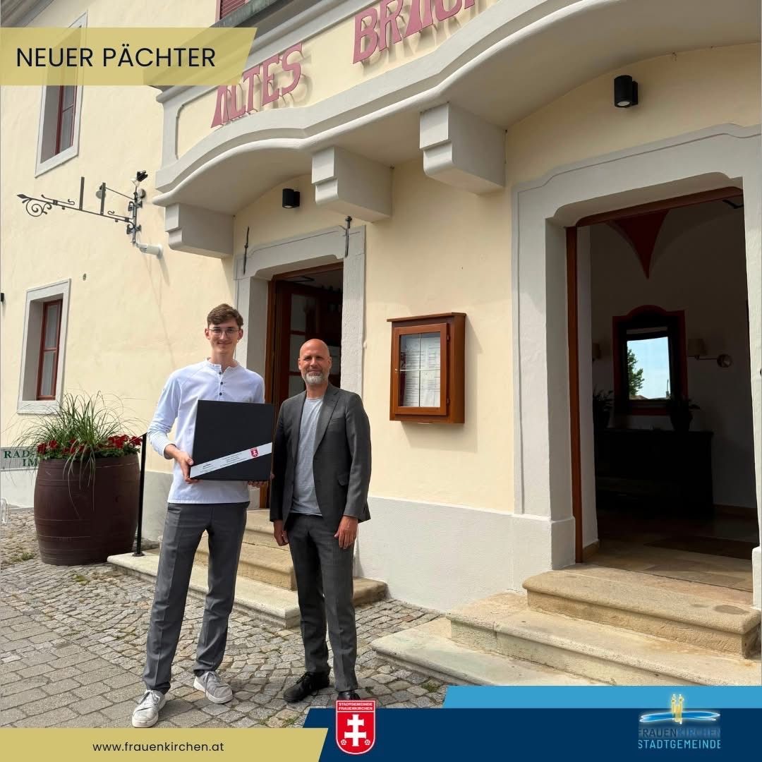 Zwei Männer stehen vor einem Gebäude mit der Aufschrift 'Alter Pächter'. Der Mann auf der linken Seite hält eine Mappe und lächelt. Der Mann auf der rechten Seite sieht in die Kamera. In der Nähe steht eine große Topfpflanze.