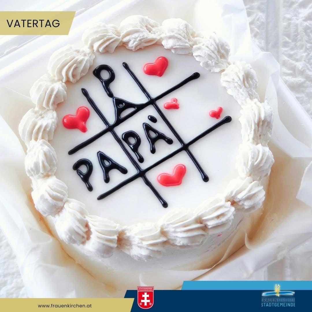 Ein Kuchen mit weißer Glasur hat ein schwarzes Gitter mit Herzen und dem Wort 'Papa' in der Mitte. Der Kuchen liegt auf einem weißen Tuch und ist in Plastik verpackt. Er ist mit 'Vatertag' beschriftet.