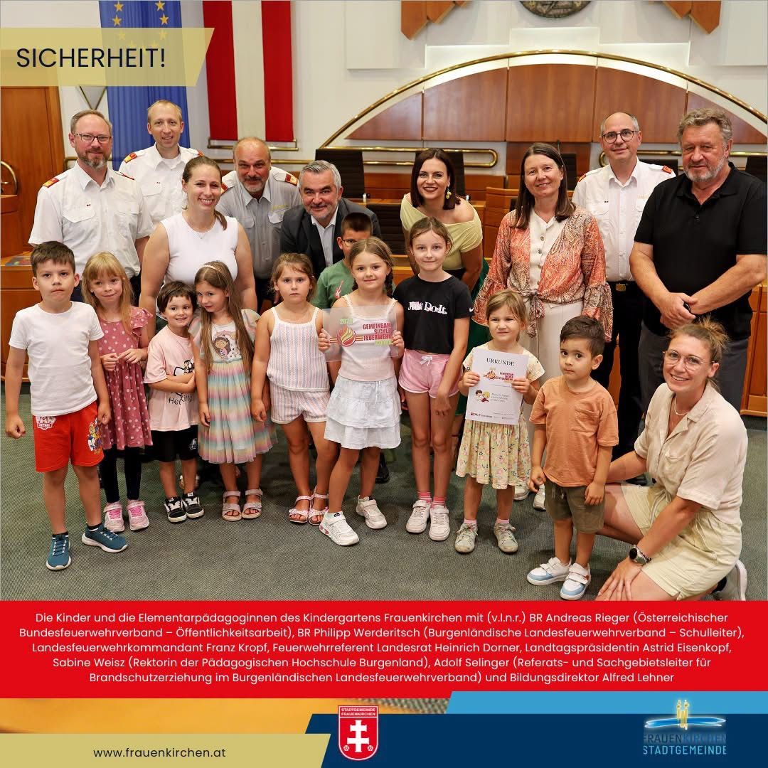 A group of children and their elementary school teachers from the Frauenkirchen Kindergarten pose for a photo with officials, including the Austrian Federal Fire Brigade Commander, in a hall.