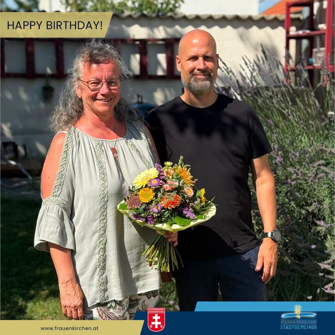 A man and a woman stand in a garden, smiling, with a bouquet of flowers. The man wears a black shirt, and the woman wears a light blouse. Behind them, a white building and a red structure are visible. The image has a birthday message at the top.
