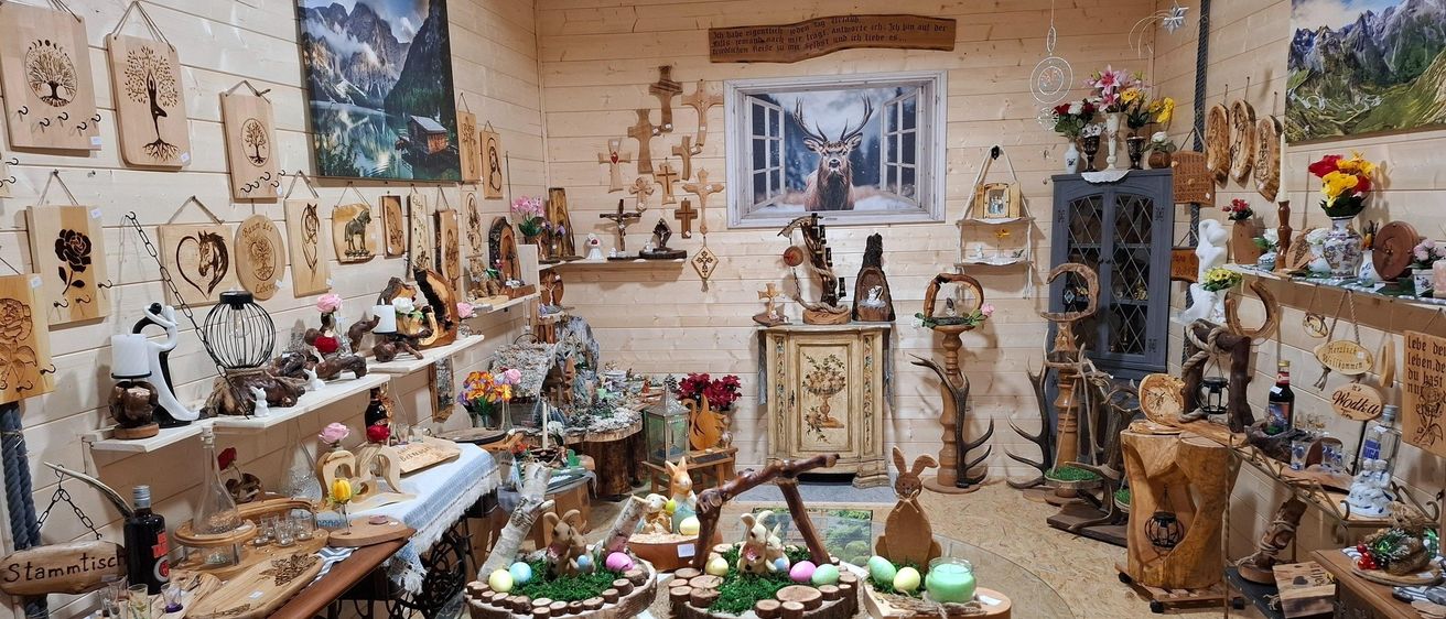 Ein Raum gefüllt mit Dekorationsartikeln, Holzschnitzereien und Osterdekorationen. Ein Tisch mit Glasplatte zeigt Ostereier und einen Kuchen. Die Wände sind mit Holztafeln und Kunstwerken geschmückt.