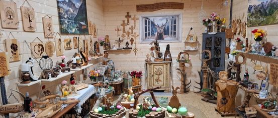 A room filled with decorative items, wooden carvings, and Easter decorations. A table with a glass top features Easter eggs and a cake. The walls are adorned with wooden plaques and artwork.