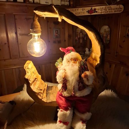 Eine hölzerne Mondsichellampe mit einer leuchtenden Glühbirne steht auf einer Holzoberfläche. Der Weihnachtsmann sitzt auf dem Halbmond und hält einen Weihnachtsbaum. Ein Pelzteppich liegt auf der Oberfläche.