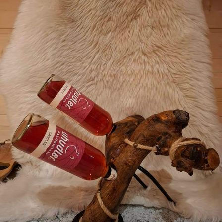 Zwei Weinflaschen werden auf einem rustikalen Holzast präsentiert, der auf einem Fellteppich ruht. Die Weinflaschen haben ein rotes Etikett mit weißem Text, der 'Primera' liest.