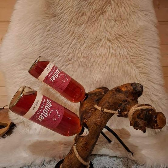 Zwei Weinflaschen werden auf einem rustikalen Holzast präsentiert, der auf einem Fellteppich ruht. Die Weinflaschen haben ein rotes Etikett mit weißem Text, der 'Primera' liest.