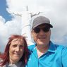 A smiling man and woman pose in front of the Christ the Redeemer statue, under a cloudy sky.