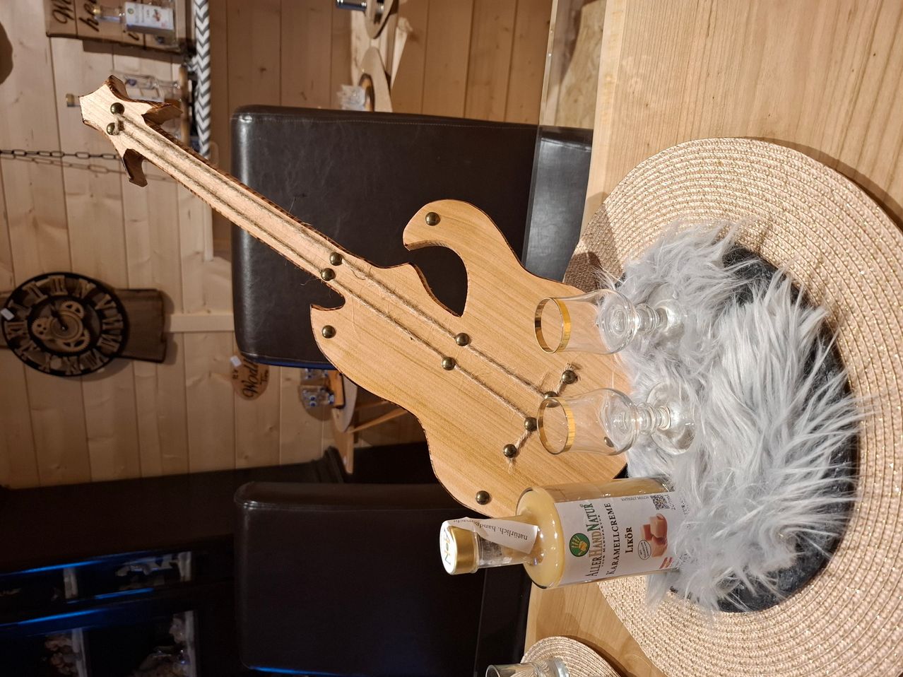 Ein Gitarrenförmiger Holzuntersetzer hält zwei Gläser und eine Flasche Karamellcreme auf einem Holztisch neben einem Hut mit Federn.