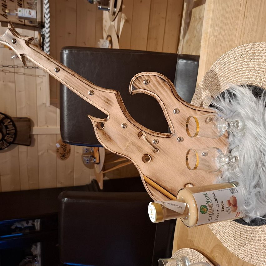 Eine hölzerne Gitarre steht auf einem Tisch, neben einem Hut und zwei Gläsern. Eine Flasche Karamellcreme ist vor der Gitarre positioniert. Dahinter ist ein schwarzer Stuhl zu sehen.