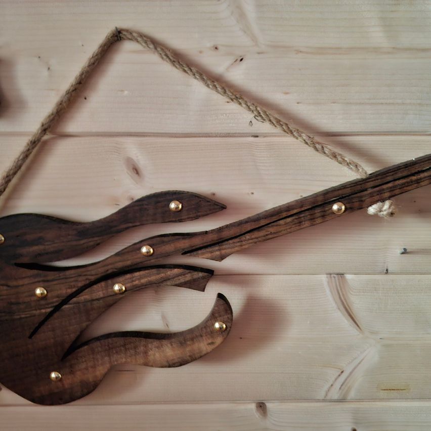 Ein Gitarren-Wandschild aus Holz mit Flammendesign hängt an einer Holzwand. Die Gitarre hat goldene Nieten und einen daran befestigten Seil.